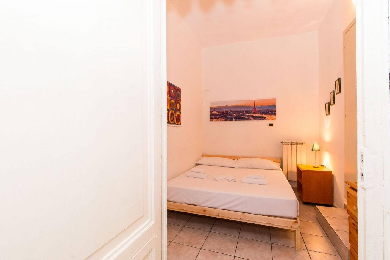 A single bed with a wooden frame and white bedding, a small bedside table with a lamp, and a wall-mounted picture of a cityscape.