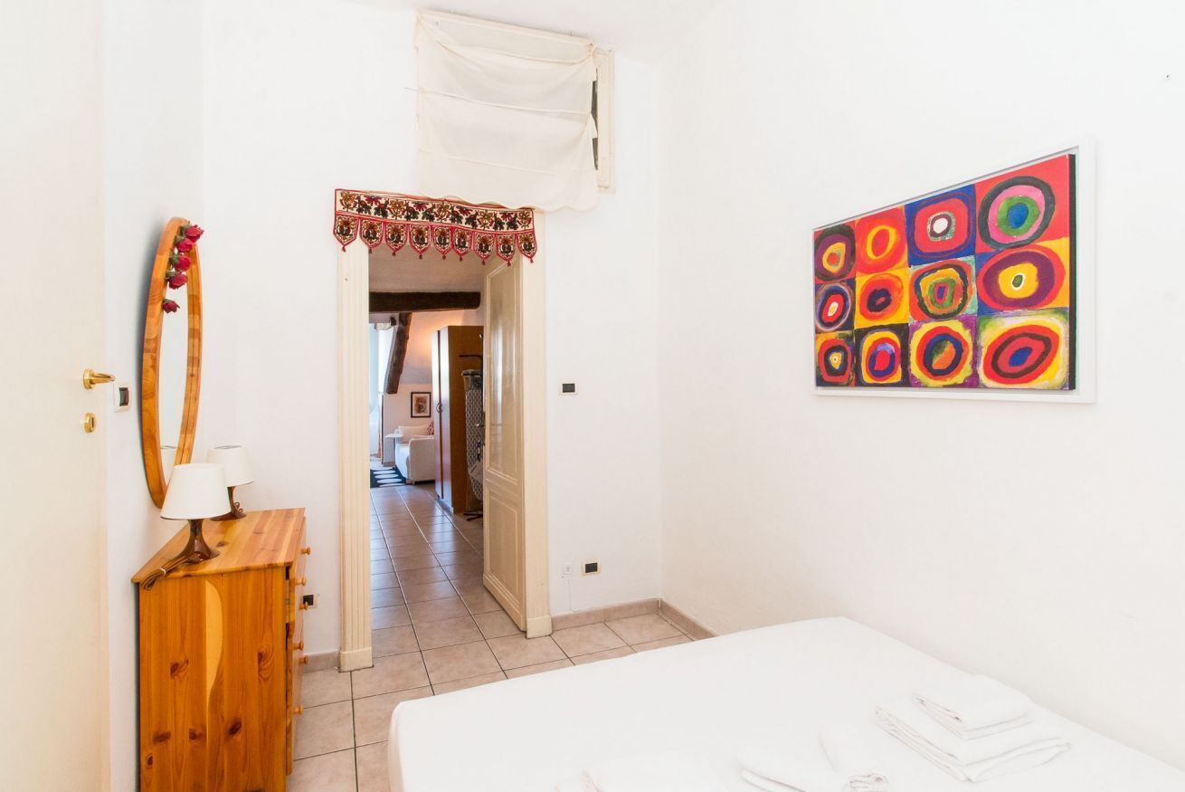 Um quarto com uma cama, penteadeira de madeira, um espelho e um quadro abstrato colorido na parede.