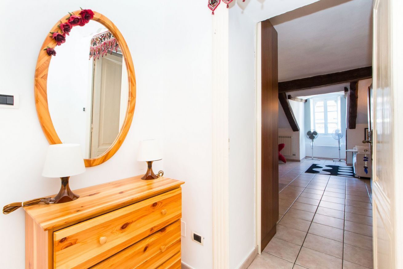A hallway with a wooden dresser, a mirror with a floral frame, and a view into a room with a window and a fan.