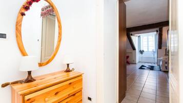 A hallway with a wooden dresser, a mirror with a floral frame, and a view into a room with a window and a fan.