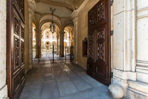 Un corridoio con porte in legno decorate e pavimento in pietra che conduce a un cortile aperto con un edificio sullo sfondo.