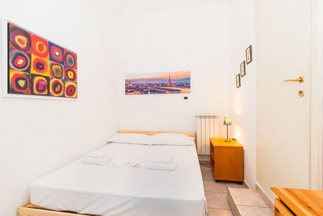 Un letto rifatto con lenzuola bianche, vista sulla città, un quadro astratto dai colori vivaci e un comodino con una lampada.
