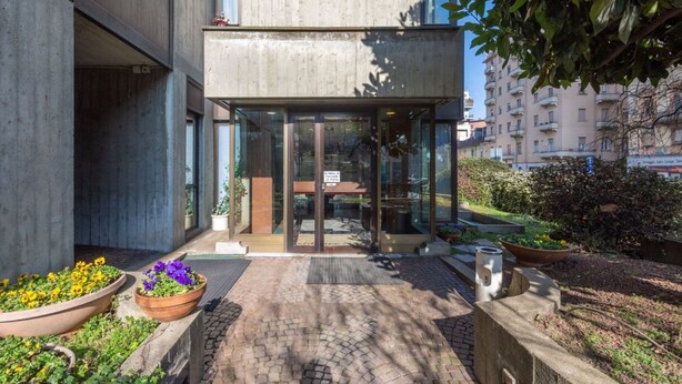 A modern building entrance with a glass door, potted plants, and a paved pathway. - Porta Nuova & Valentino Apartment (Turin)