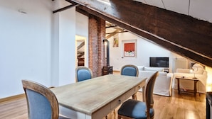 Un espace repas avec une table et des chaises en bois, une colonne en briques et un escalier menant à un salon équipé d’un téléviseur et d’un canapé.