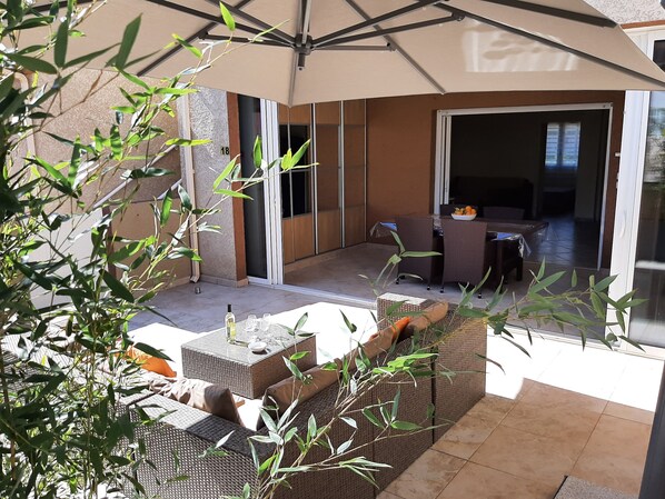 Un patio al aire libre con mobiliario de mimbre, una mesa con un cuenco de fruta y una puerta corrediza de vidrio que da a un espacio interior.