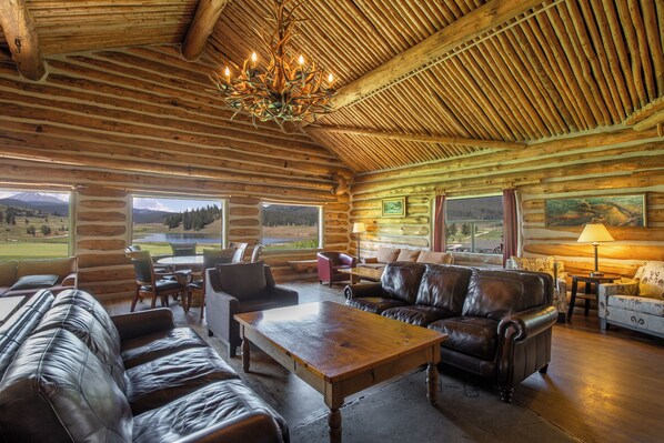 A log cabin interior with a large wooden ceiling, leather sofas, a wooden coffee table, and a view of a mountainous landscape through large windows. - 63 Forget Me Not Lane by Summit County Mountain Retreats (Keystone)
