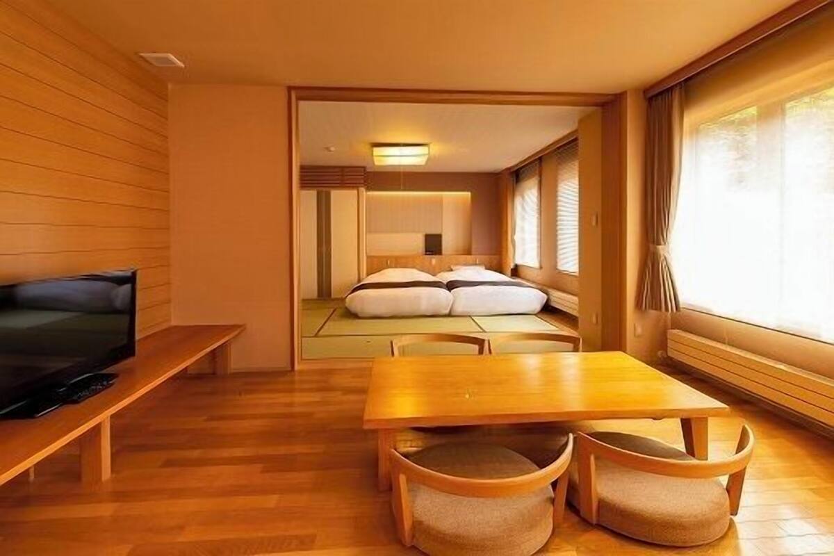 Een kleine kamer met een bed, een televisie op een standaard, een houten tafel en een raam met gordijnen.