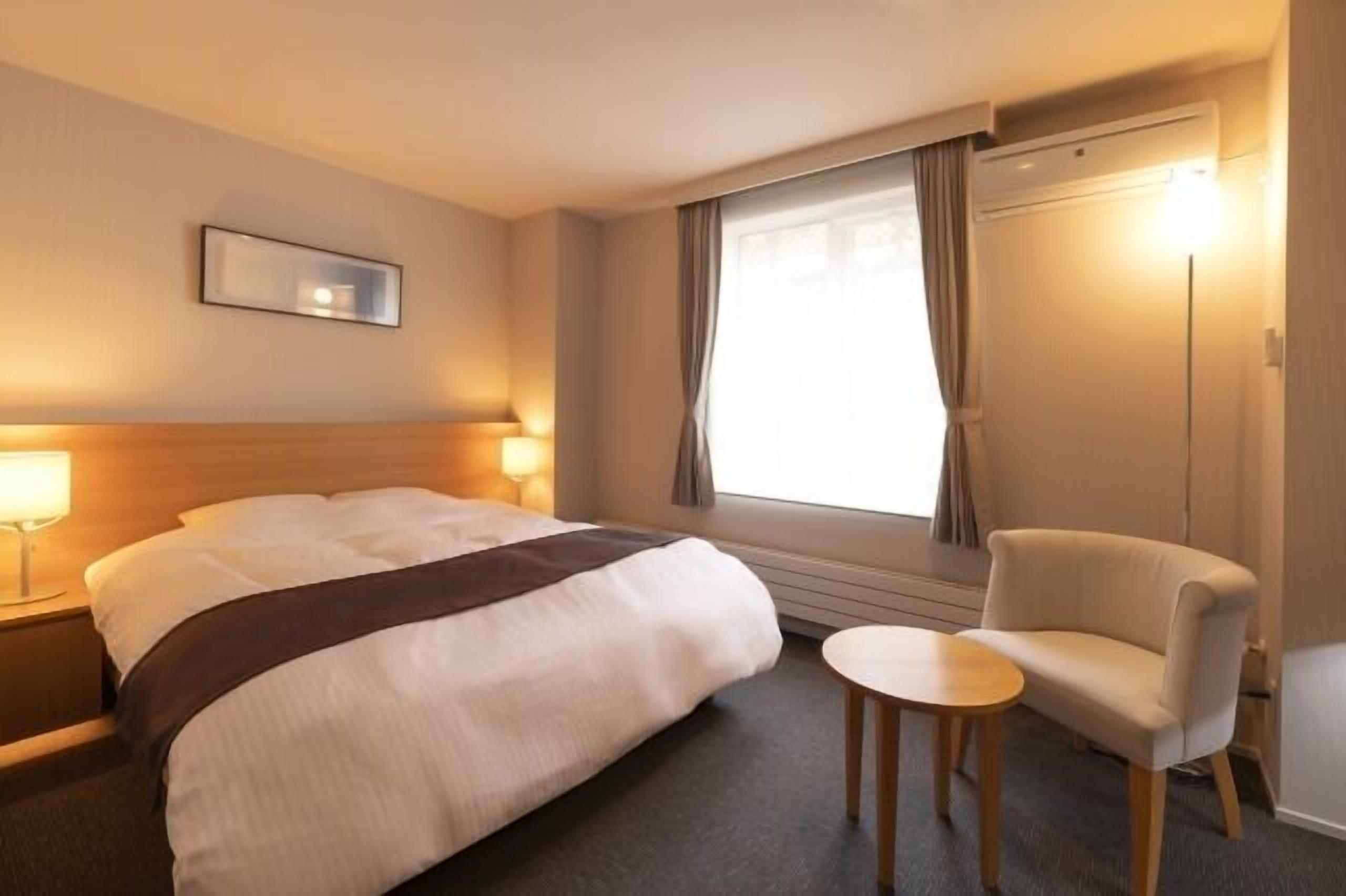 Habitación de hotel con cama, mesita de noche, lámpara, silla y ventana con cortinas.
