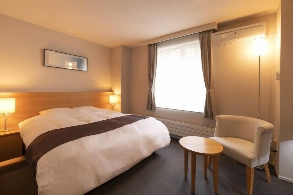 Een hotelkamer met een bed, nachtkastje, lamp, stoel en raam met gordijnen.