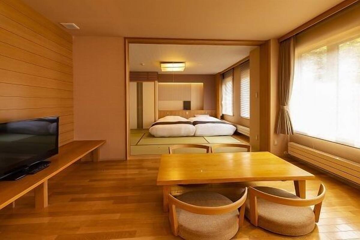Een compacte hotelkamer met houten vloer, een televisie op een bankje, een bed met kussens, een klein tafeltje en een raam met gordijnen.