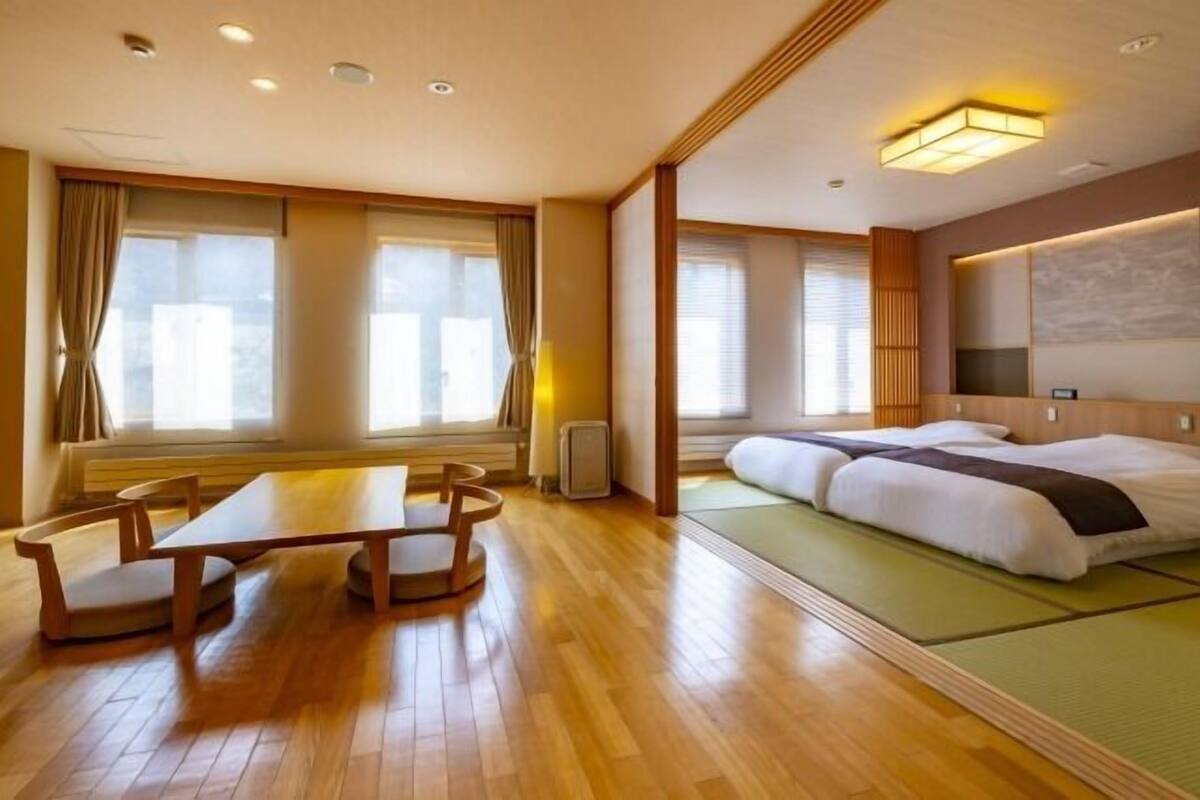 Een hotelkamer met een houten vloer, een bed met groene matrassen, een tafel met stoelen en grote ramen voorzien van gordijnen.