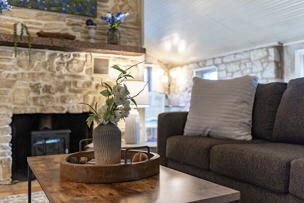 A cozy living room with a stone fireplace, a wooden coffee table with a vase of flowers, and a grey sofa with cushions.