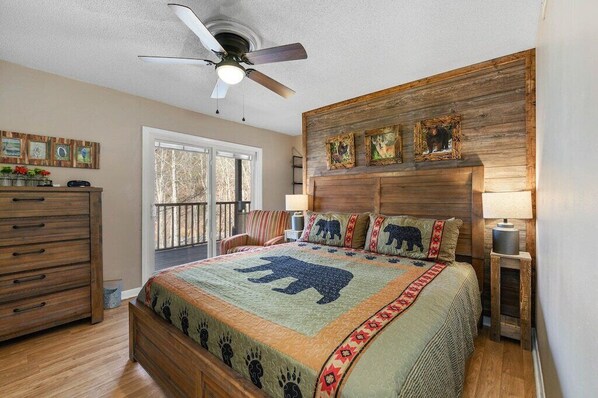 Un dormitorio con cabecera de madera, cama con colcha estampada con motivos animales, ventilador de techo y ventana con vista a árboles.