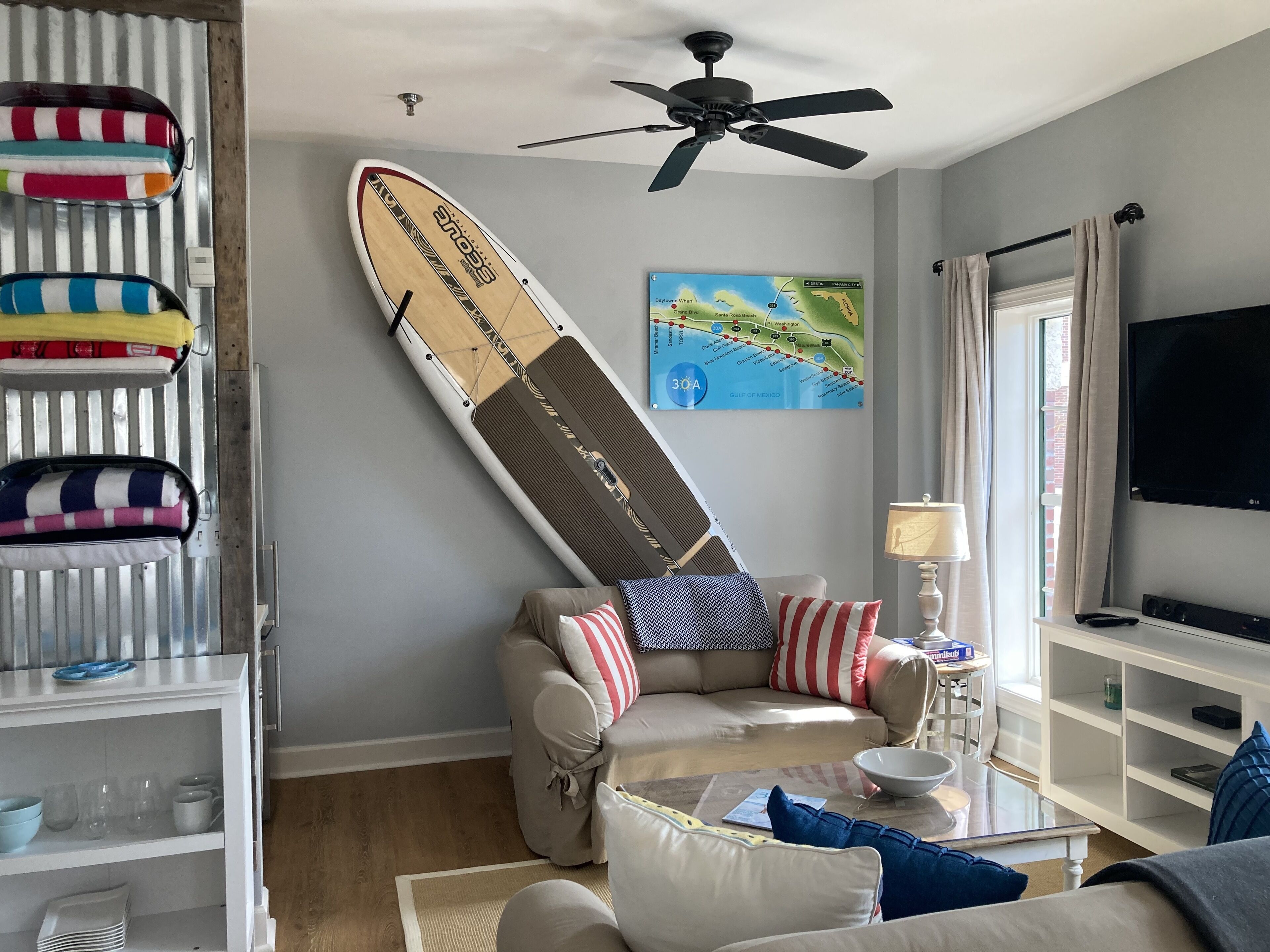 Un salon avec un canapé, une table basse, un téléviseur, un ventilateur de plafond, une carte accrochée au mur et une planche de surf appuyée contre le mur.
