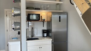 Cocina compacta con refrigerador de acero inoxidable, microondas y cafetera sobre la encimera. Estantes arriba de la encimera con platos, vasos y un objeto decorativo.