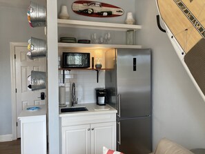 Cocina compacta con refrigerador de acero inoxidable, microondas y cafetera sobre la encimera. Estantes arriba de la encimera con platos, vasos y un objeto decorativo.