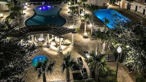 Vista nocturna de un resort con dos piscinas, un área de patio techada y sillas de descanso.