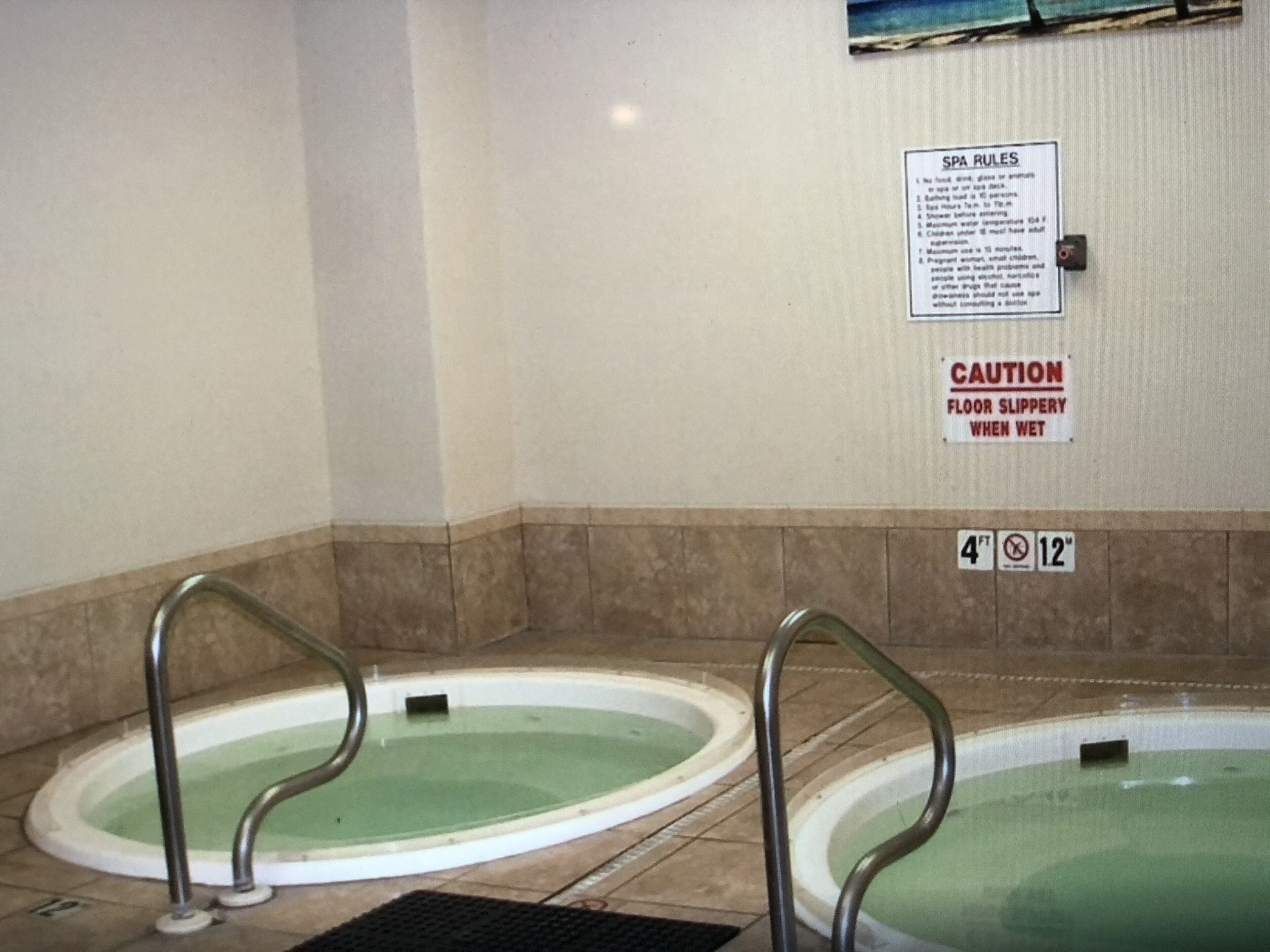 Zona de spa con dos jacuzzis, señal de precaución por suelo resbaladizo cuando está mojado y un cartel con las normas del spa.