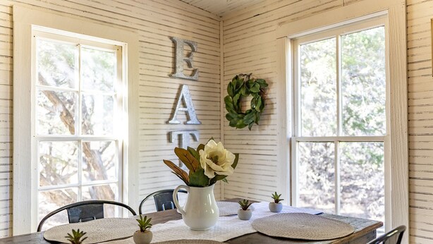 Un comedor con una mesa de madera, individuales, pequeñas macetas y una jarra blanca con flores.