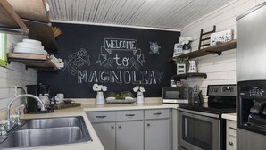 Una cocina con un panel de pizarra que muestra “BIENVENIDOS A MAGNOLIA”, electrodomésticos de acero inoxidable y una cafetera.