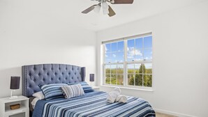 A bedroom with a tufted headboard, a large window with a view, a bedside table with a lamp, and a striped bedspread.