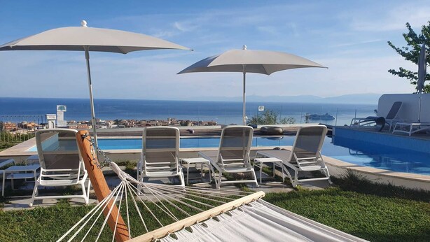 A hammock with white cushions, a poolside lounge area with white chairs and umbrellas, and a view of the ocean and distant buildings. - Appartamenti Borgo Panoramico N.6 (Tropea)
