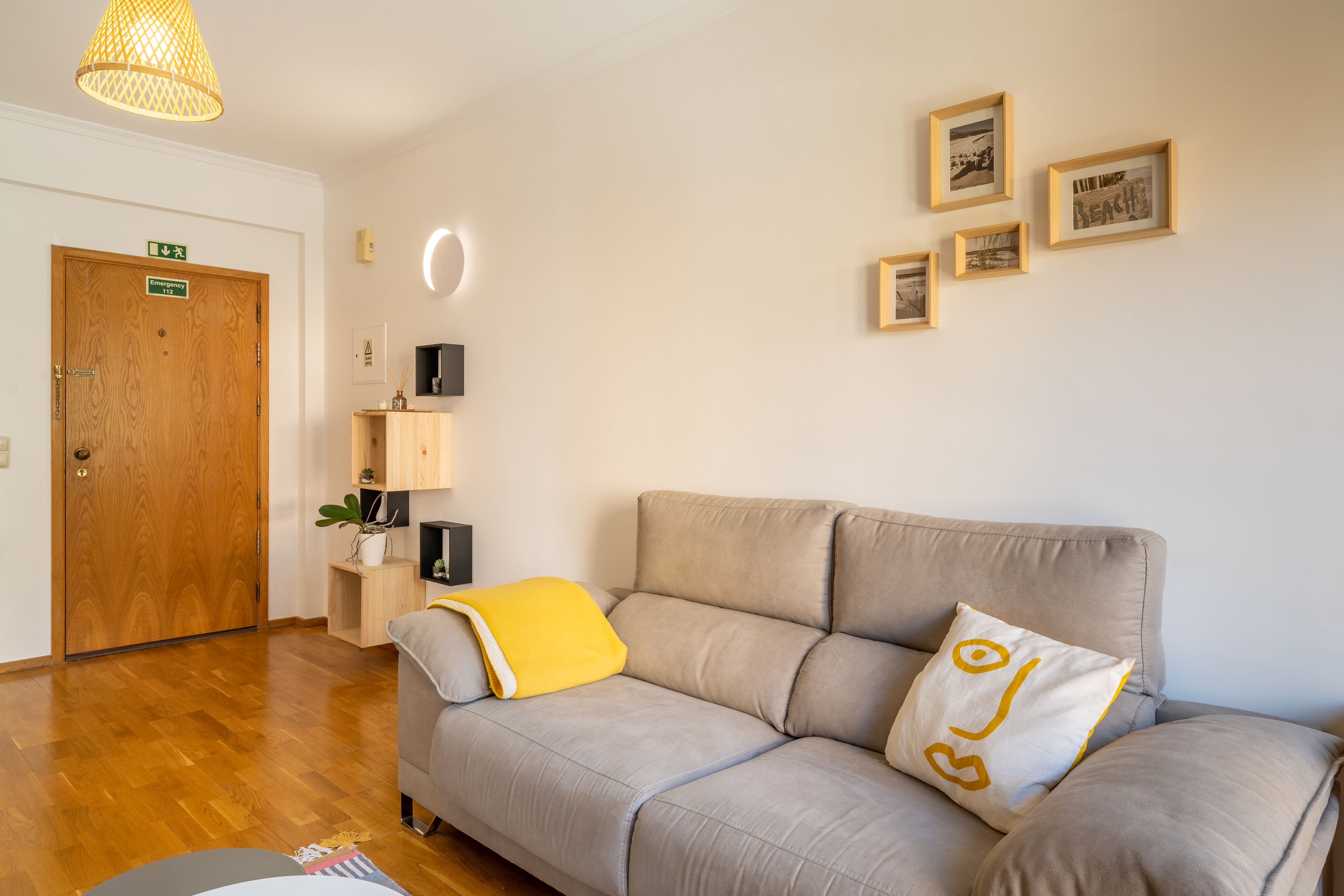 Un salon moderne avec un canapé gris, un coussin jaune et une porte en bois portant un panneau indiquant la sortie de secours.