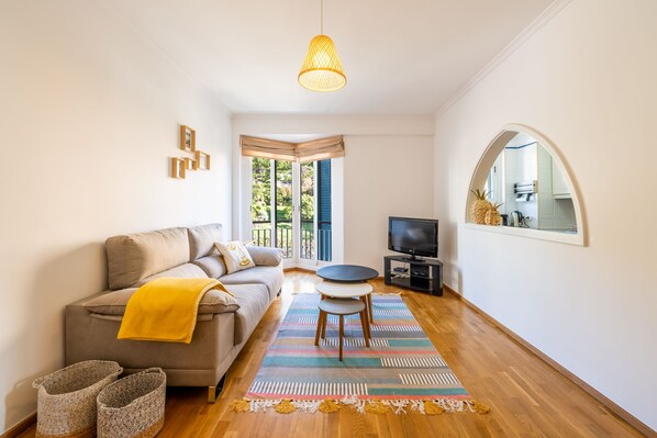 A living room with a sofa, a coffee table, a TV, and a window with a view of greenery.