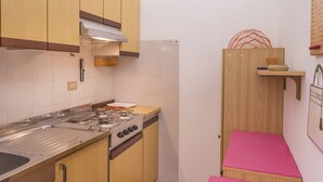 A compact kitchen with wooden cabinets, a gas stove, and a small wooden shelf. - Lignano Sea & Relax Apartment (Lignano Sabbiadoro)