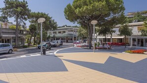 A public square with a central open area, surrounded by buildings, trees, and a few parked cars. - Lignano Sea & Relax Apartment (Lignano Sabbiadoro)