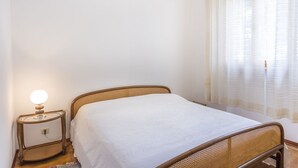 A single bed with a wooden headboard, a bedside table with a lamp, and sheer curtains on a window. - Lignano Sea & Relax Apartment (Lignano Sabbiadoro)