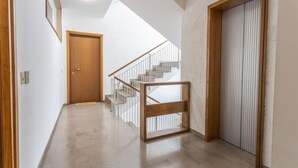 Een gang met een houten deur, een wenteltrap met een metalen leuning, en een groot raam dat voor natuurlijk licht zorgt.