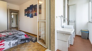 A bedroom with a bed, a large window, a bathroom with a sink and washing machine, and a cityscape artwork on the wall. - Zia Dina Apartment (Pasian di Prato)