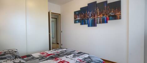 A bedroom with a bed, a wardrobe, and a cityscape painting on the wall.