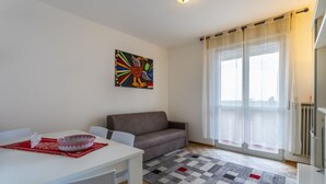 A modern living room with a sofa, a dining table with a bowl, a colorful abstract painting, and a large window with sheer curtains. - Zia Dina Apartment (Pasian di Prato)