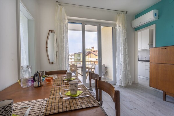 A dining area with a wooden table set for four, a kitchen with white cabinets, and a view through a glass door to the outside. - Casa di Annika - Vista Laguna e Parcheggio privato (Grado)