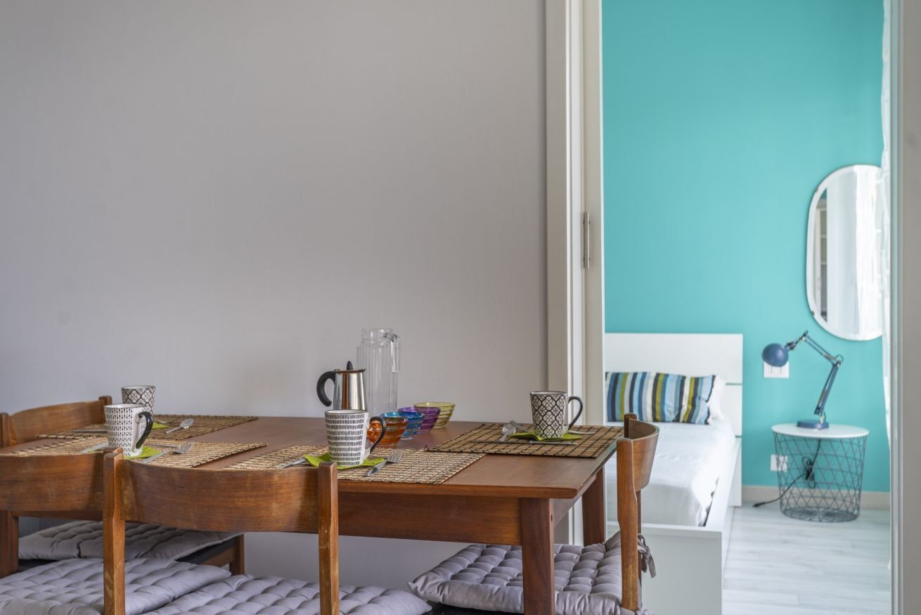 A dining area with a wooden table set for four, surrounded by cushioned chairs. A bed with a striped pillow is visible in the background.
