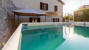 A swimming pool with a stone wall and a white building with shutters in the background. - Borgo Santa Lucia Apartment with Private Parking & Garden (Gubbio)