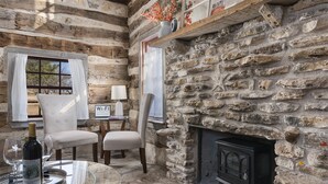 Interior rústico de una cabaña de troncos con chimenea de piedra, una mesa de comedor con sillas y una botella de vino sobre la mesa.