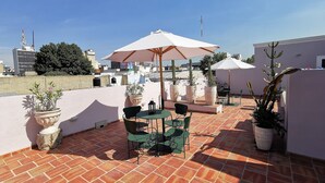 A rooftop terrace with a table and chairs under a large umbrella, surrounded by potted plants and a clear view of the city skyline.