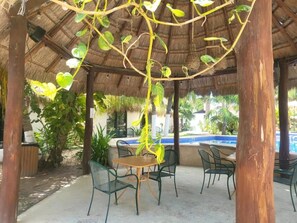 Un área de estar al aire libre con techo de paja, rodeada de follaje y una piscina al fondo.