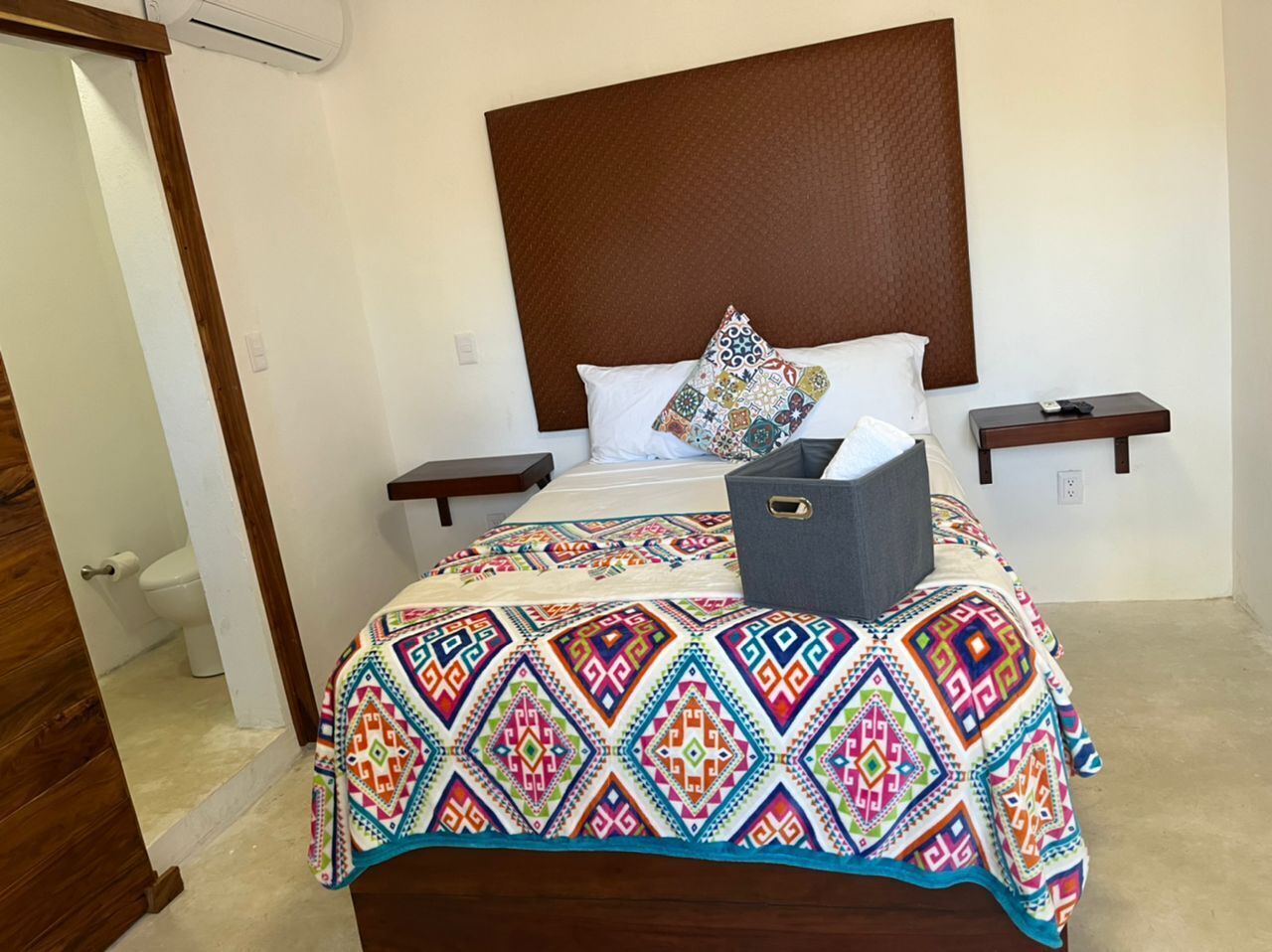 A bed with a colorful geometric-patterned bedspread, a wooden headboard, two bedside tables, and a gray storage box on the bed.