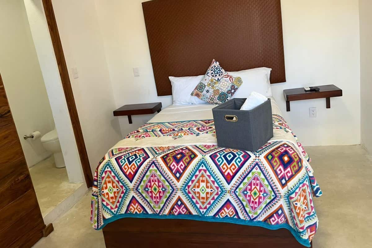 Un lit avec une couvre-lit aux motifs géométriques colorés, une tête de lit en bois, deux tables de chevet et une boîte de rangement grise posée sur le lit.