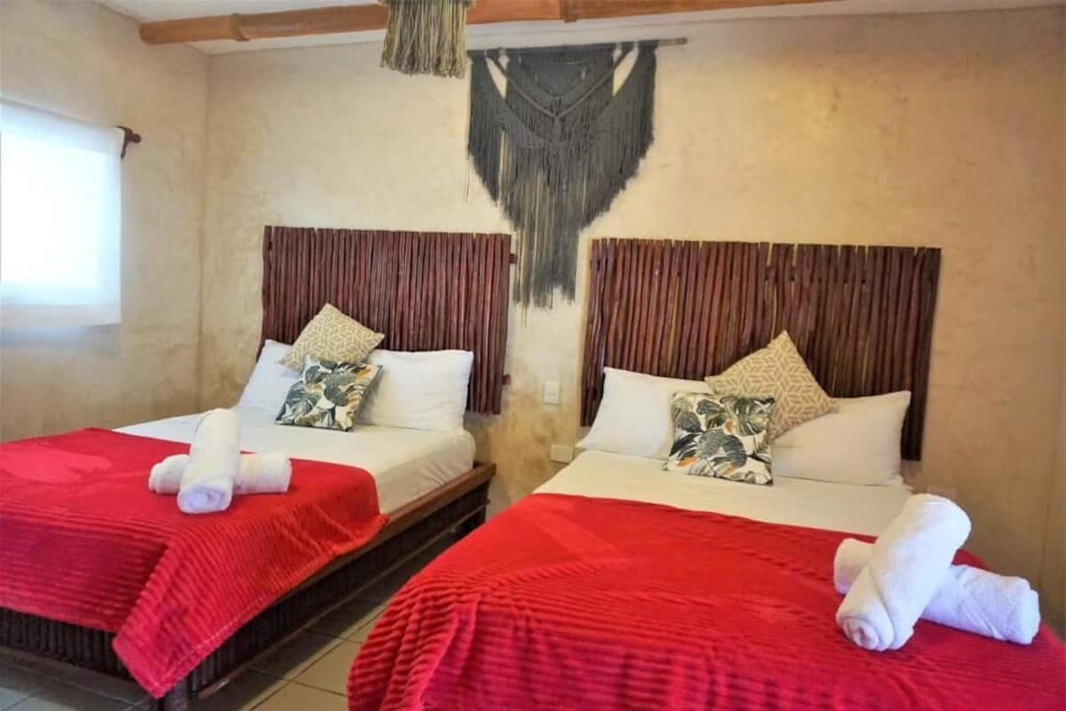 Deux lits avec une literie rouge et des oreillers blancs, une décoration murale en macramé suspendue et une fenêtre avec des rideaux.