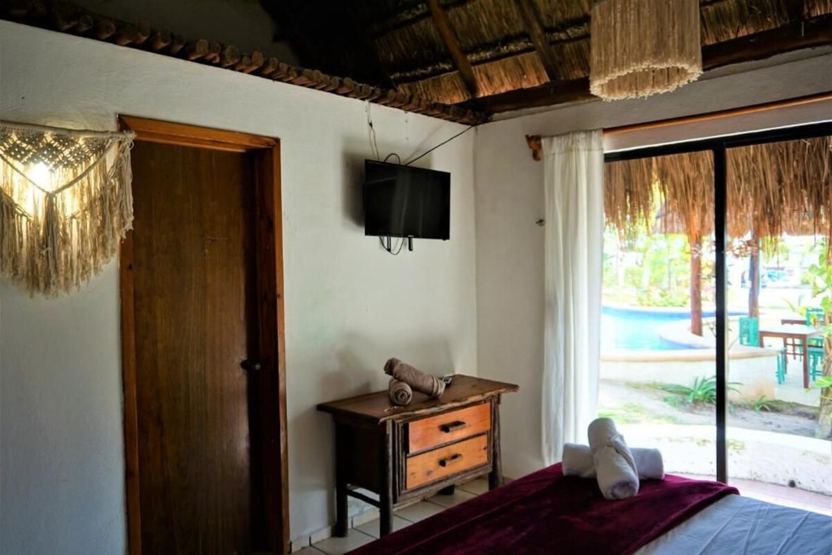 A room with a bed, a wooden nightstand, a television, and a view of an outdoor area with a pool.
