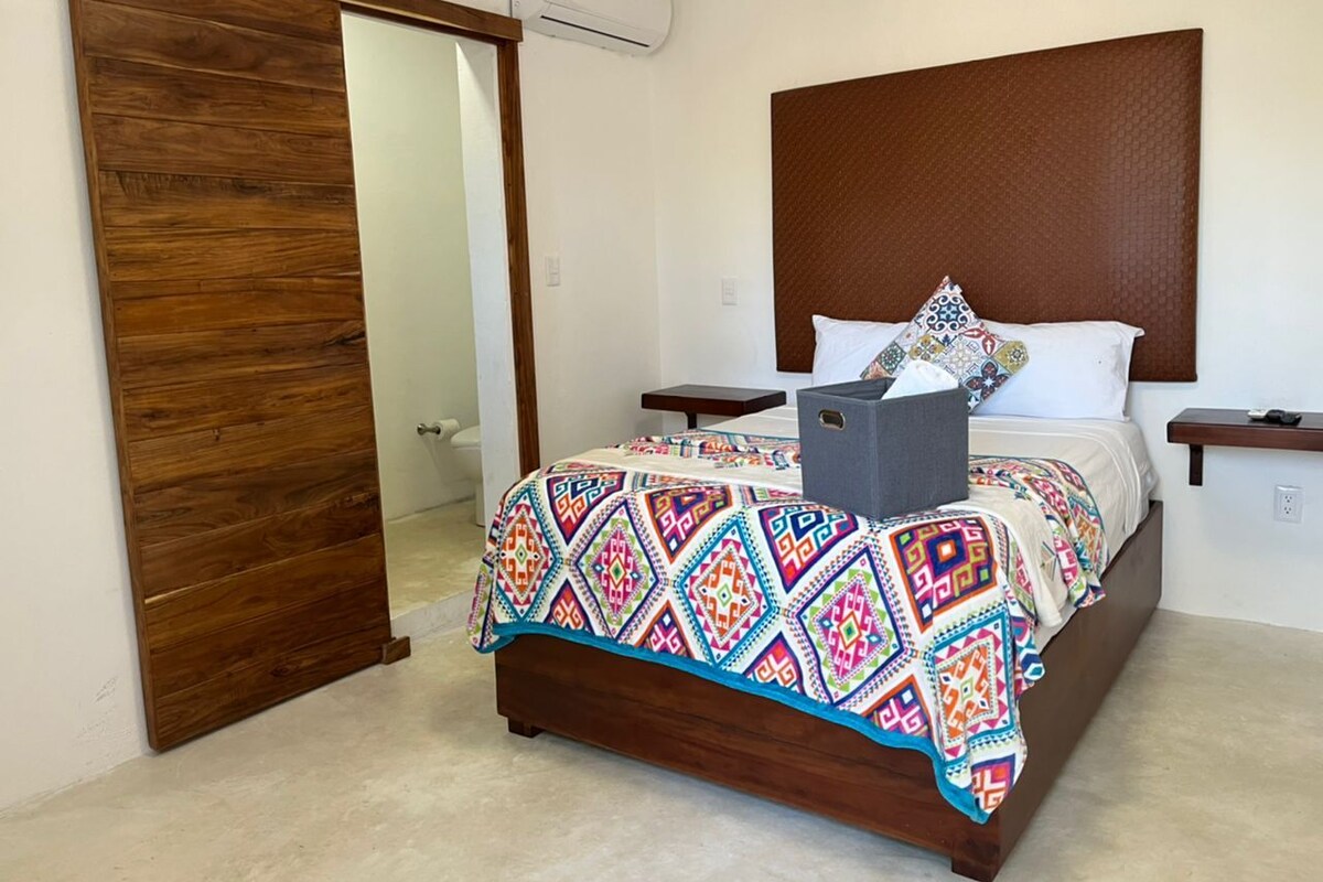 A bedroom with a wooden bed frame, a colorful quilt, a gray storage box, and a wooden headboard.
