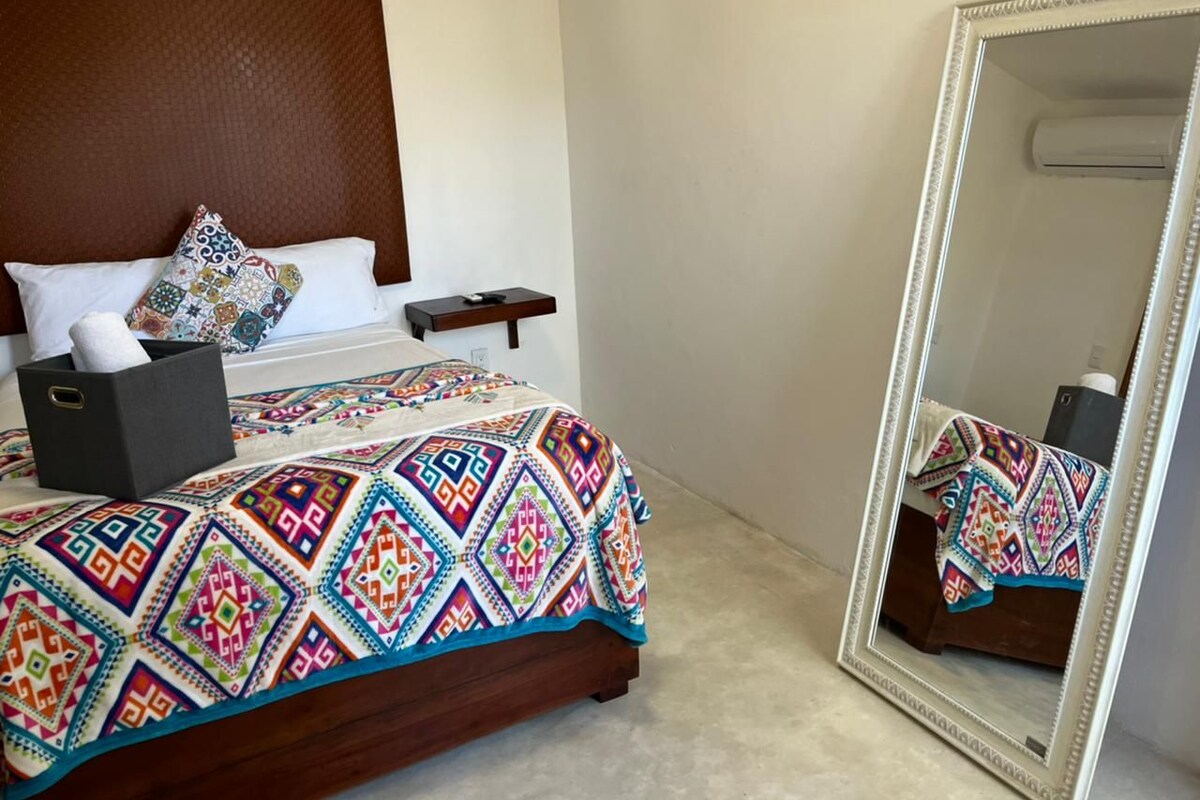 A hotel room with a wooden headboard, a colorful bedspread, a bedside table with a lamp, and a large mirror.
