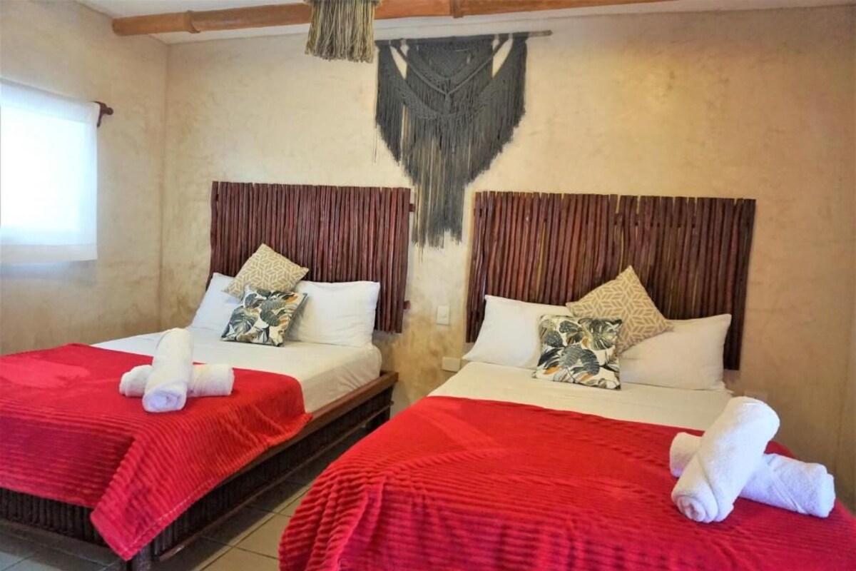 Two beds with red bedding and white pillows, a hanging macrame wall decoration, and a window with curtains.