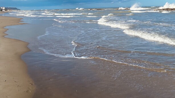 Una playa con olas acercándose a la orilla, un cielo despejado y formaciones rocosas a lo lejos.