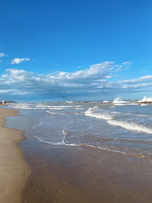 Une plage où les vagues se brisent près du rivage, un ciel dégagé et, au loin, des formations rocheuses.